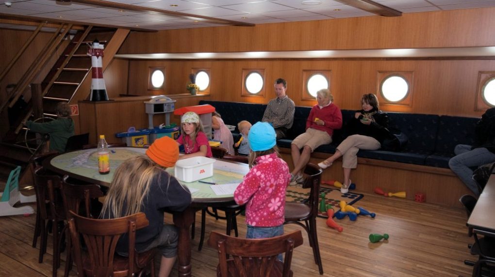 Children and adults in a cozy ship interior with tables, chairs, and portholes.