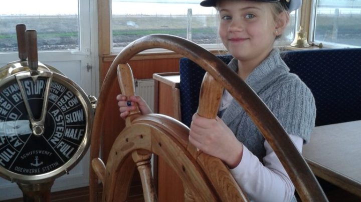 Child in captain's hat steering a ship's wheel inside a cabin.