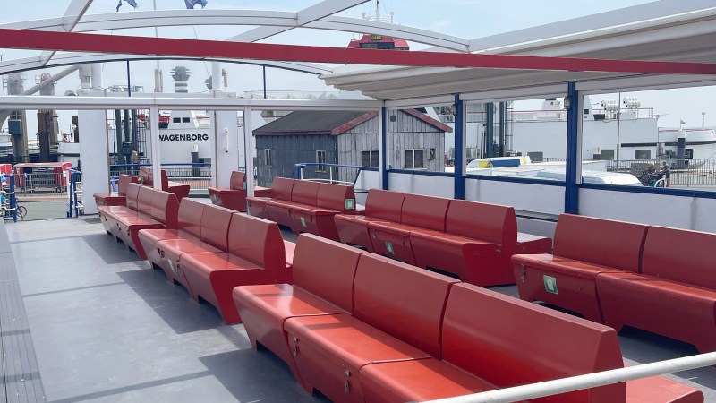 Empty ferry deck with red benches and open-air roof structure, docked at port.
