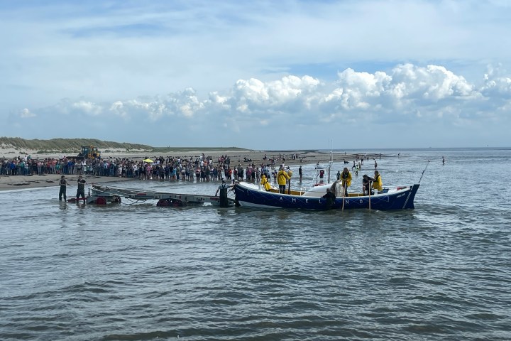 Rescue boat with crew in water, crowd on beach under blue sky.