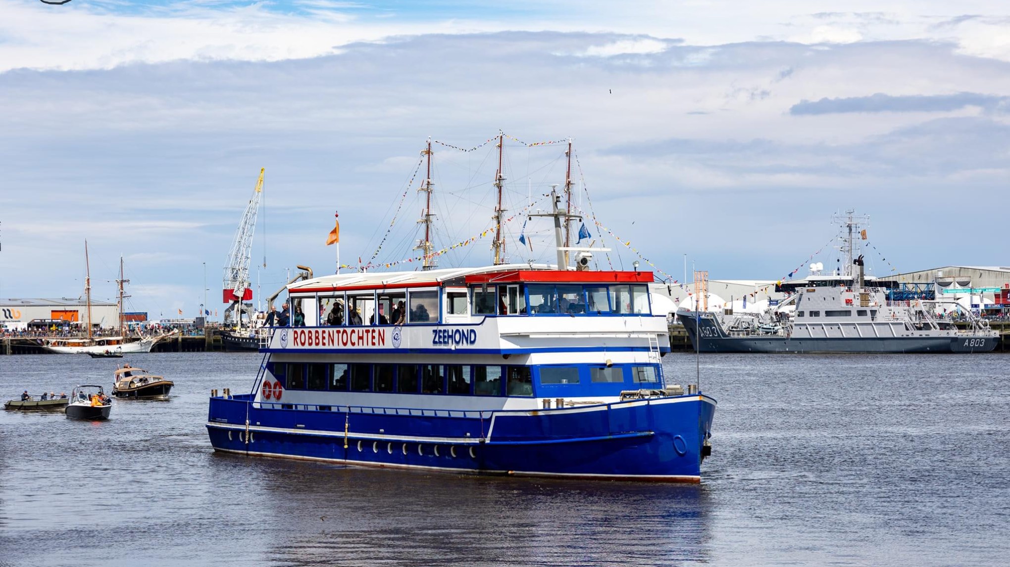 Blue tour boat in harbor with other boats and ships in the background under a cloudy sky.
