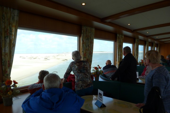 People on a ferry observing the sea and sandy shores through large windows.