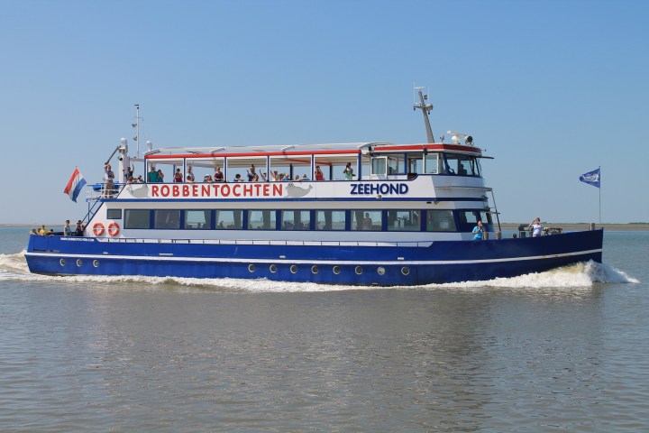 Blue and white tour boat named 'Zeehond' on water, with people on deck and clear sky.
