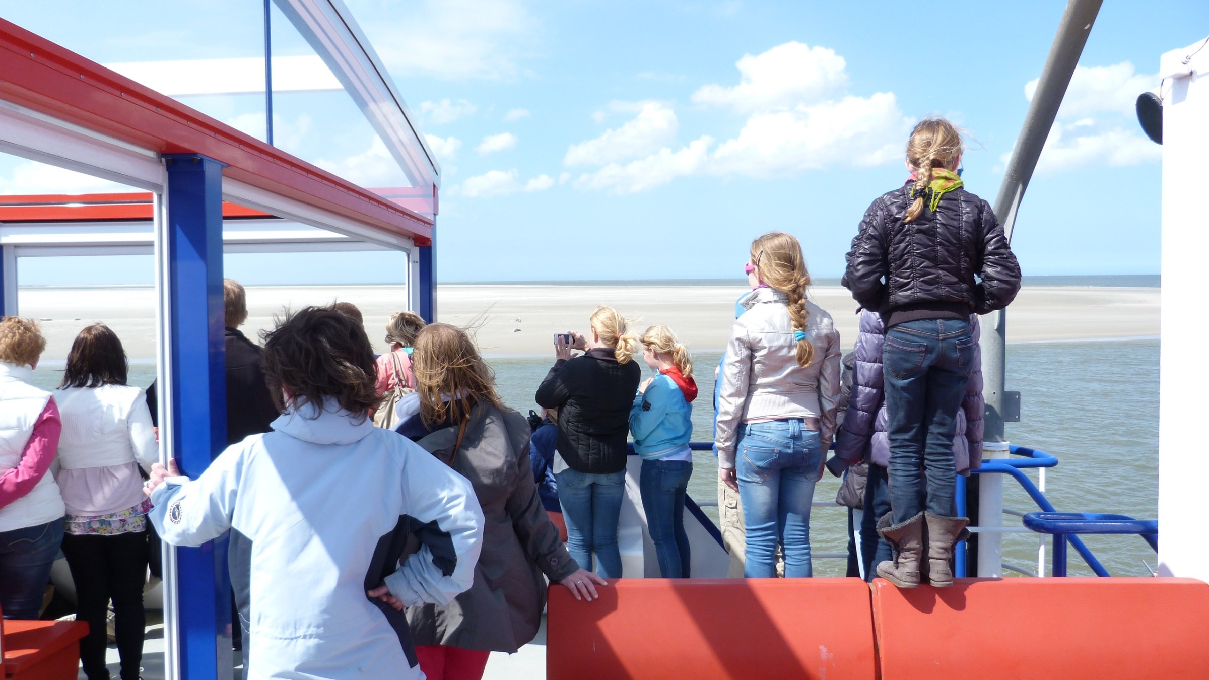 People standing on a boat deck, gazing at the sea and distant sandy shore.