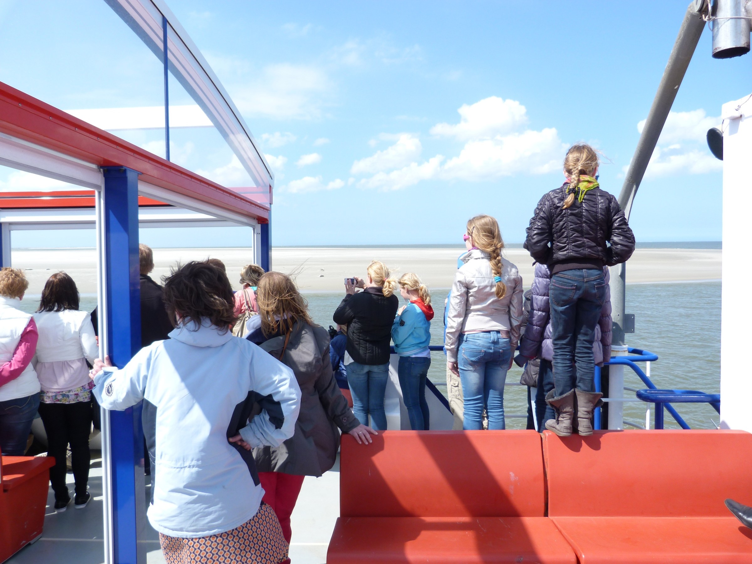 People standing on a boat deck, gazing at the sea and distant sandy shore.
